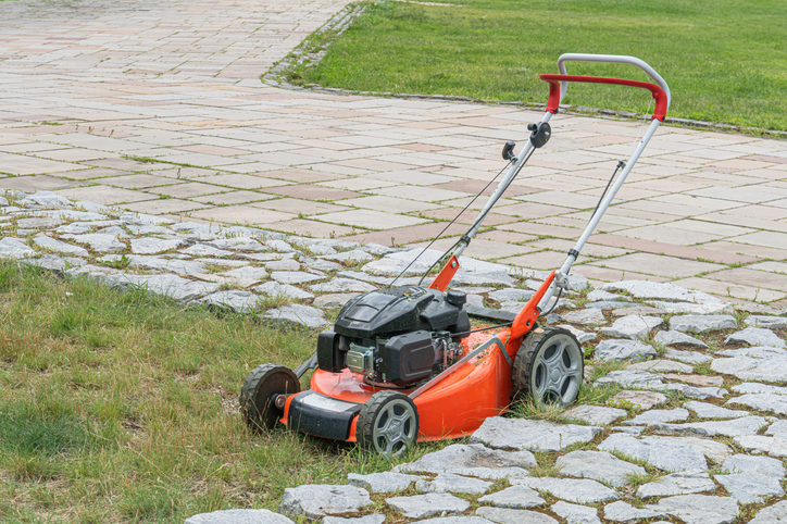 Red lawn mower with large wheels on grassy area, possibly a park with concrete path No branding or texts visible Natural lighting, no shadows suggesting harsh sunlight or artificial sources