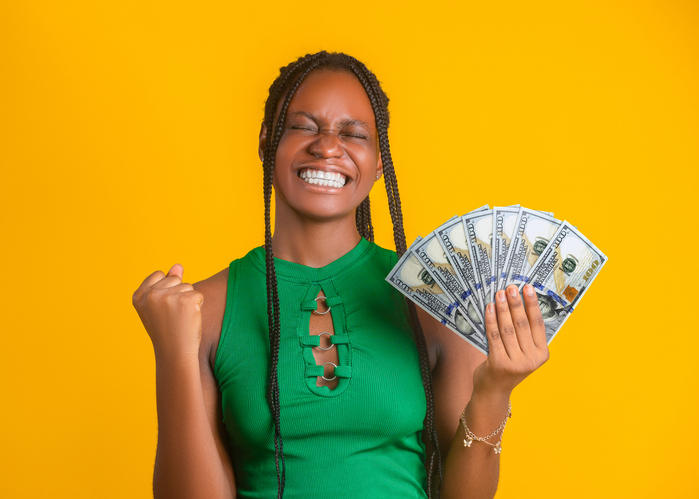 A joyful woman in a green top celebrates, holding a fan of US $100 dollar banknotes against a vibrant yellow background. Her eyes are closed in a wide, triumphant smile with a clinched fist.