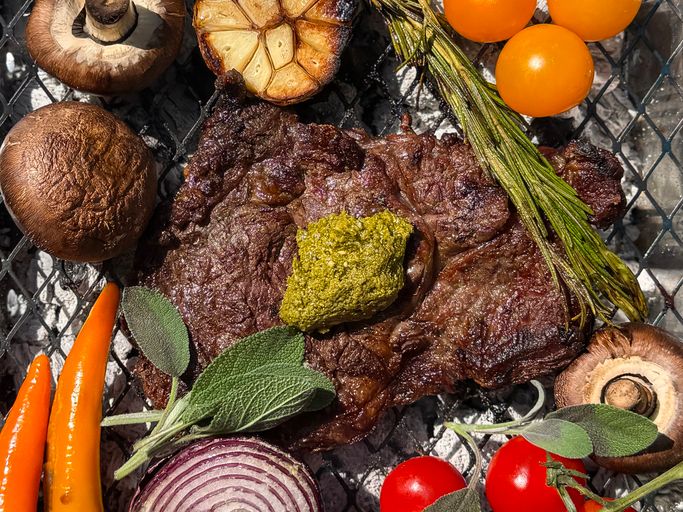 Image of Charcoal barbecue grilled aberdeen angus ribeye beef steak cooked medium rare with green pesto herbs, fresh tomatoes, garlic
