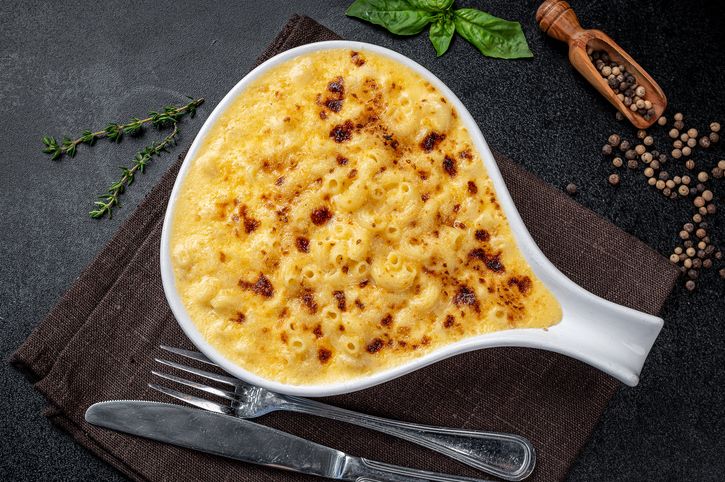 Creamy macaroni and cheese in a bowl. A bowl filled with baked macaroni and cheese sits on a dark surface beside herbs and a spoon.