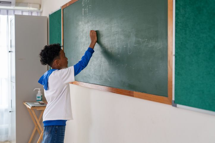 African American boy student writing math equations on green chalkboard in classroom at school
