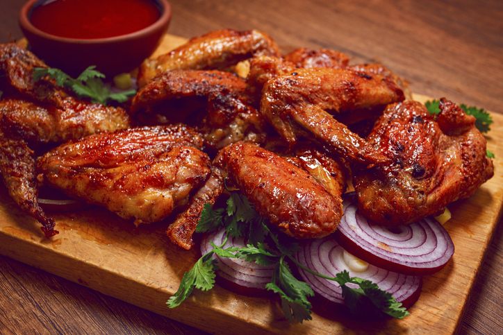 Cooked chicken wings lay on a wooden board. There are slices of onion and cilantro around the wings. A bowl of red sauce is next to the wings for dipping